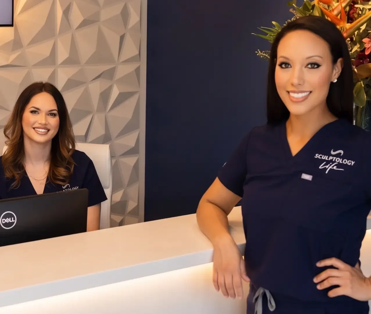 Two women in navy scrubs at a reception desk; one stands smiling, the other sits with a Dell laptop.