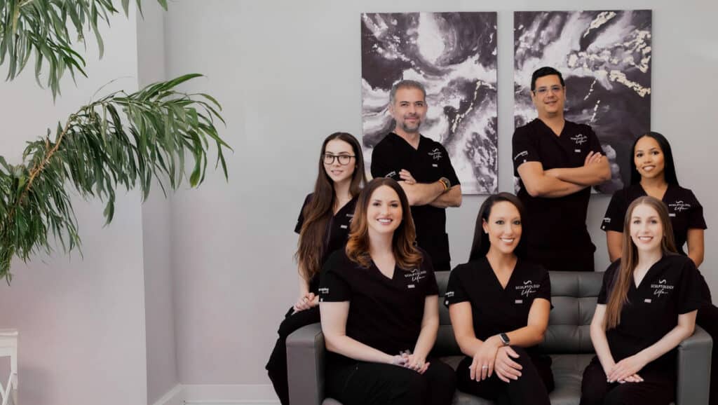Group of six medical professionals in black scrubs posing in a modern clinic lounge with a large plant and abstract wall art.